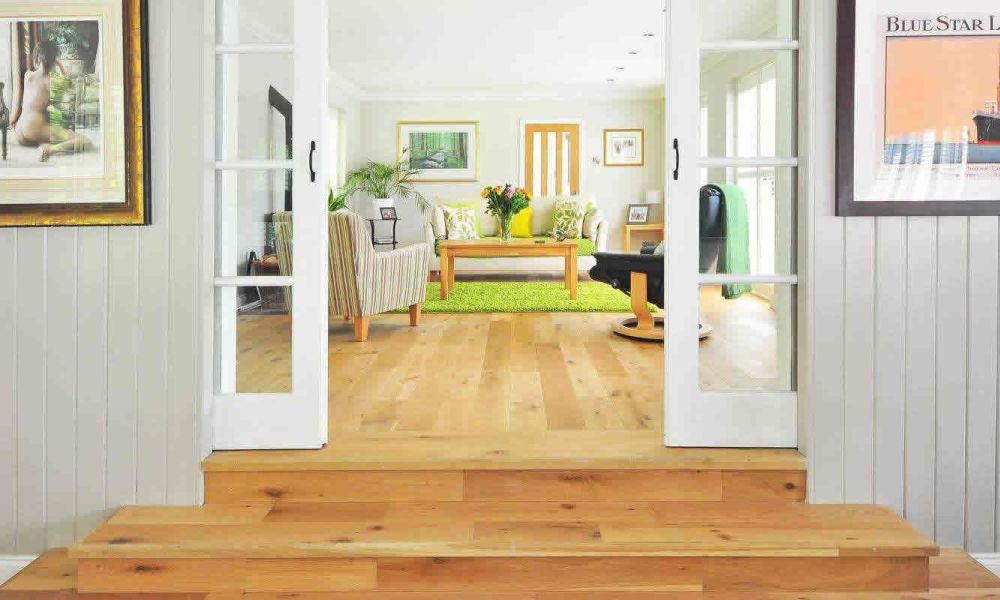 <span class="uk-h4">A view into a bright living room with a large window, a green rug, and comfortable seating, seen through open French doors from a hallway with art on the walls.</span>