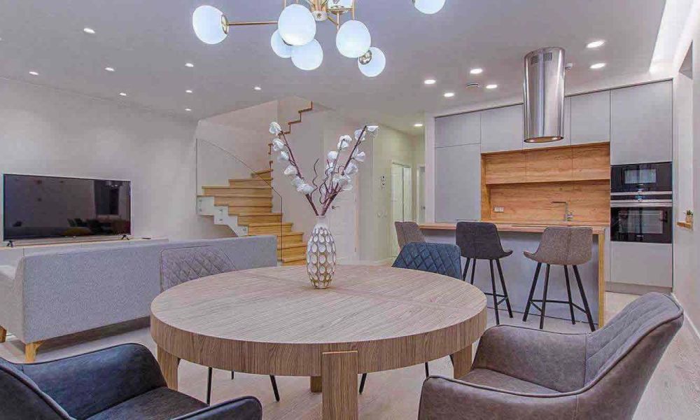 <span class="uk-h4">An open-concept living and dining area with a round dining table, gray chairs, a staircase, and a modern kitchen, showcasing a blend of functionality and style.</span>