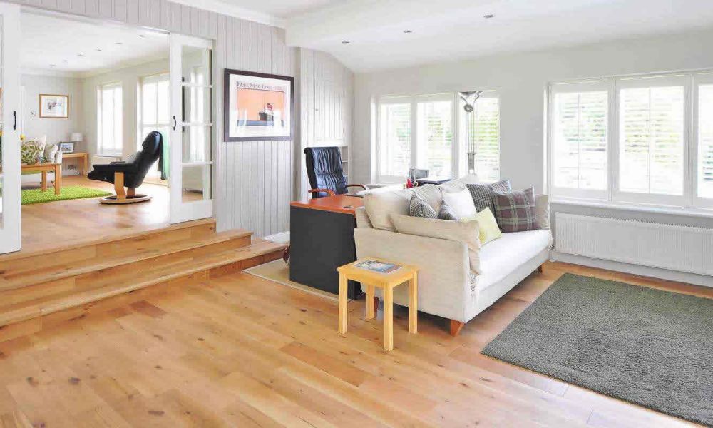 <span class="uk-h4">A spacious living area with light wooden floors, a comfortable sofa, a black desk, and large windows with white blinds, offering a bright and inviting atmosphere.</span>