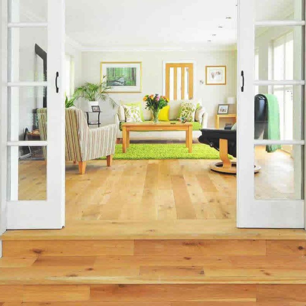 <span class="uk-h4">A view into a bright living room with a large window, a green rug, and comfortable seating, seen through open French doors from a hallway with art on the walls.</span>