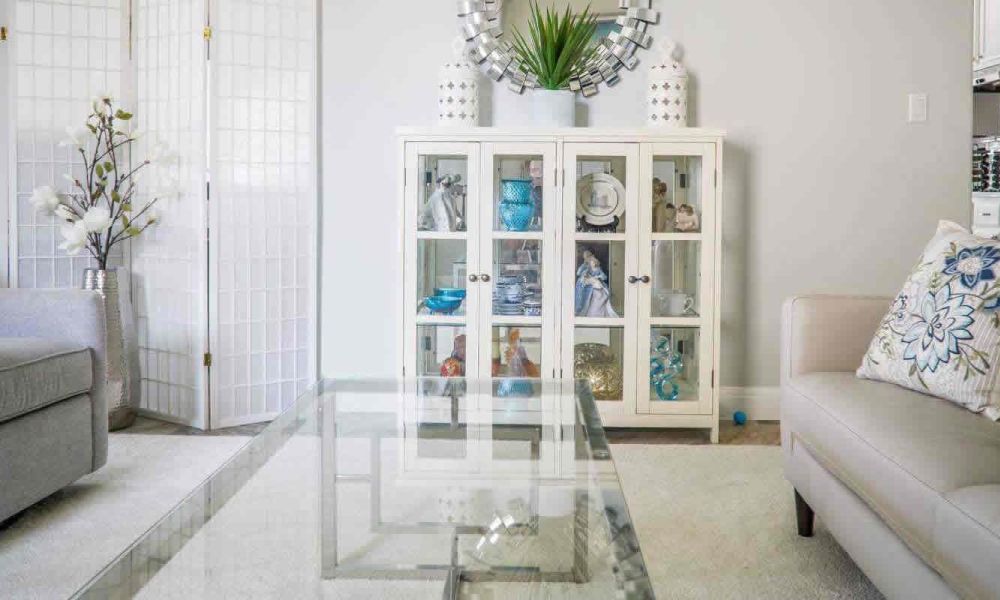 <span class="uk-h4">A living room with a glass coffee table, a white display cabinet with decorative items, and a comfortable sofa, exuding a fresh and elegant feel.</span>