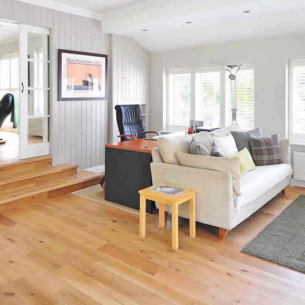 <span class="uk-h4">A spacious living area with light wooden floors, a comfortable sofa, a black desk, and large windows with white blinds, offering a bright and inviting atmosphere.</span>