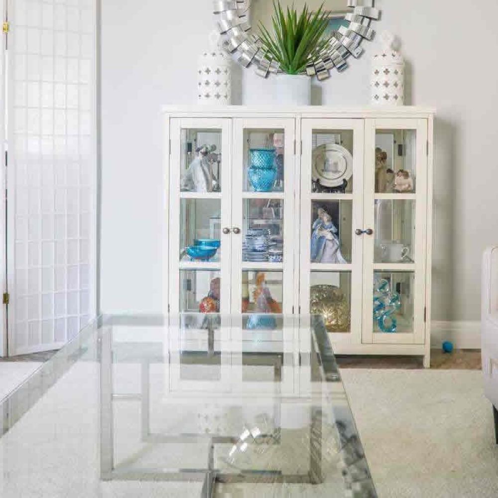 <span class="uk-h4">A living room with a glass coffee table, a white display cabinet with decorative items, and a comfortable sofa, exuding a fresh and elegant feel.</span>