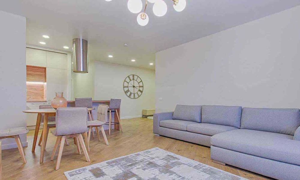 <span class="uk-h4">A modern living room with a gray sectional sofa, a dining area with a round table and chairs, and stylish lighting fixtures, creating a cozy and elegant ambiance.</span>